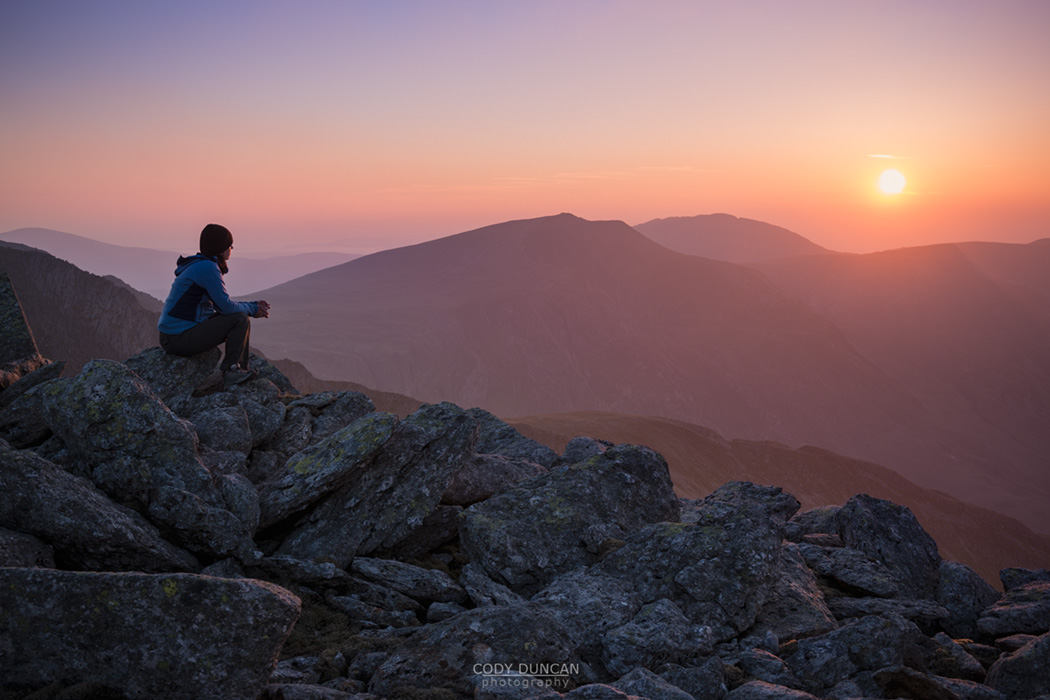 Female hiker watches sunset over mountains from summit of Glyder Fach, Snowdonia national park, Wales