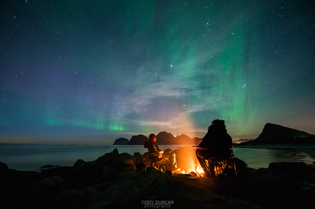 Sitting around an campfire with northern lights - aurora borealis in the sky overhead,Storsandnes, Flakstadøy, Lofoten Islands, Norwya